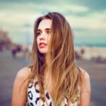 Beautiful young girl walking on city ocean beach boardwalk at sunset time and wind blowing up her hair. Woman looking to the side outdoors.