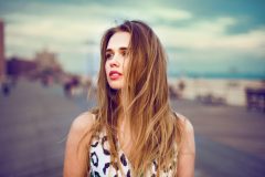 Beautiful young girl walking on city ocean beach boardwalk at sunset time and wind blowing up her hair. Woman looking to the side outdoors.